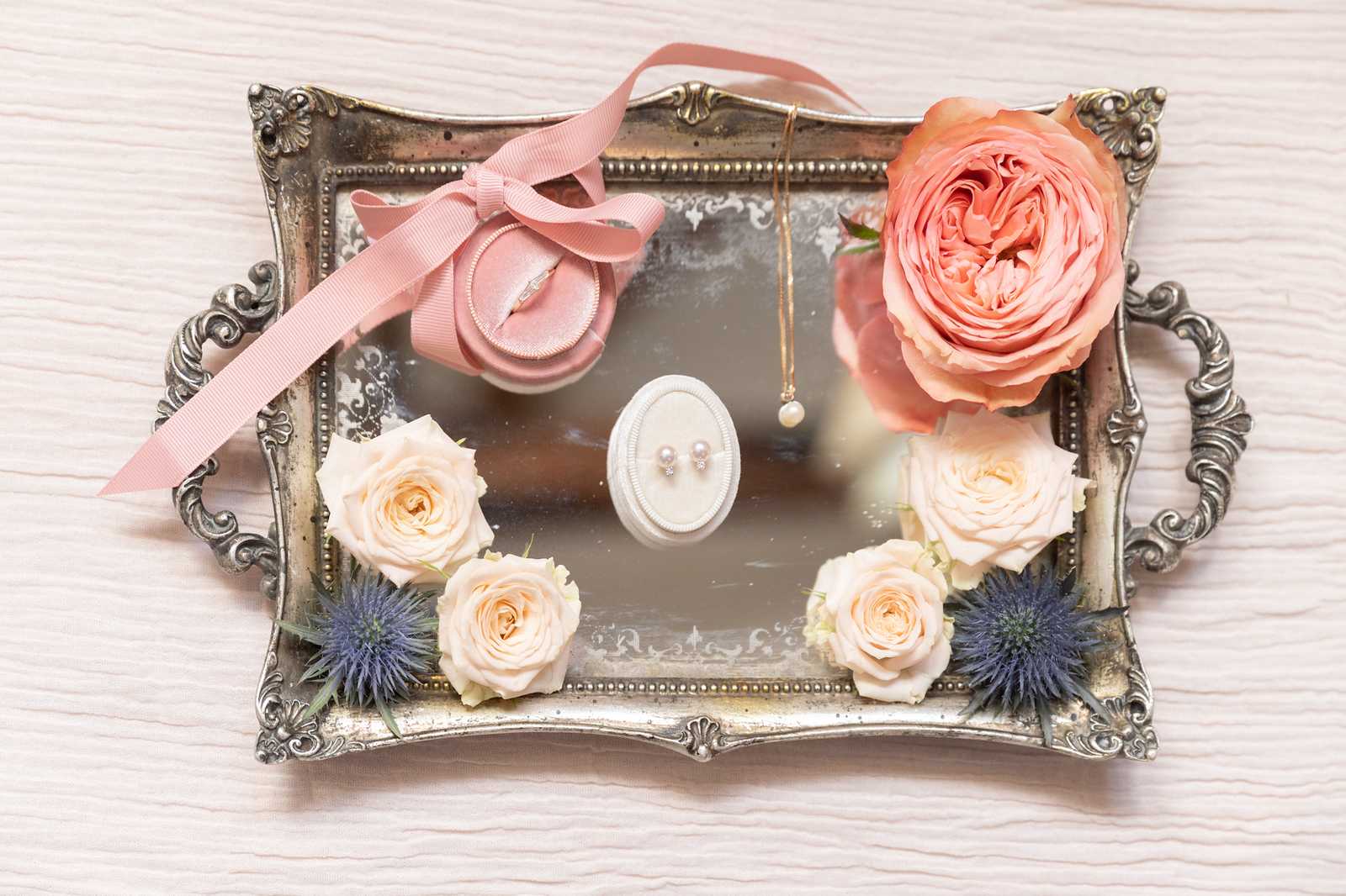 A flat lay detail shot of bridal accessories arranged on an ornate silver mirrored tray with decorative handles and embossed border. The tray holds a dusty pink velvet ring box tied with a grosgrain ribbon bow containing a diamond engagement ring, a white oval jewelry stand displaying pearl stud earrings, and a delicate gold chain necklace with a single pearl pendant. Floral styling includes one large coral garden rose in the upper right corner, four small cream spray roses, and several blue-purple thistle heads arranged around the lower portion of the tray. The overall color palette combines coral, cream, dusty pink, and steel blue in a romantic, vintage-inspired styling.