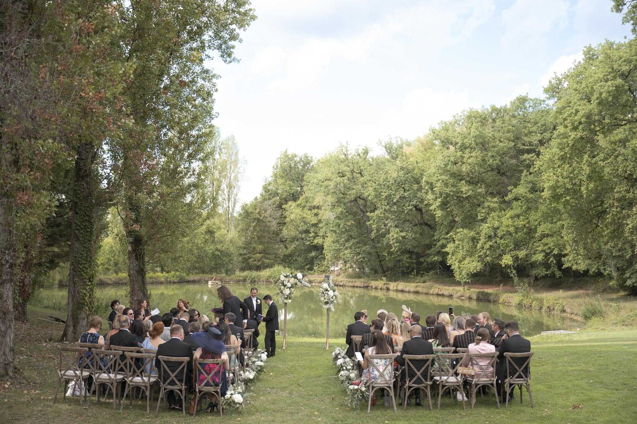 An outdoor wedding ceremony taking place on a lawn directly beside a calm river or pond, with a dense treeline as the backdrop. Approximately 60–80 guests are seated in two sections on wooden cross-back chairs, facing a simple tall floral arch decorated with white blooms and greenery at the altar. The aisle is lined with low white floral arrangements and foliage. Two figures in dark suits stand at the altar alongside an officiant, suggesting a same-sex or mixed-gender ceremony. The overall decor palette is white and green with a natural, garden-style aesthetic. The shot is a wide, slightly elevated angle capturing the full ceremony setup and surrounding landscape.