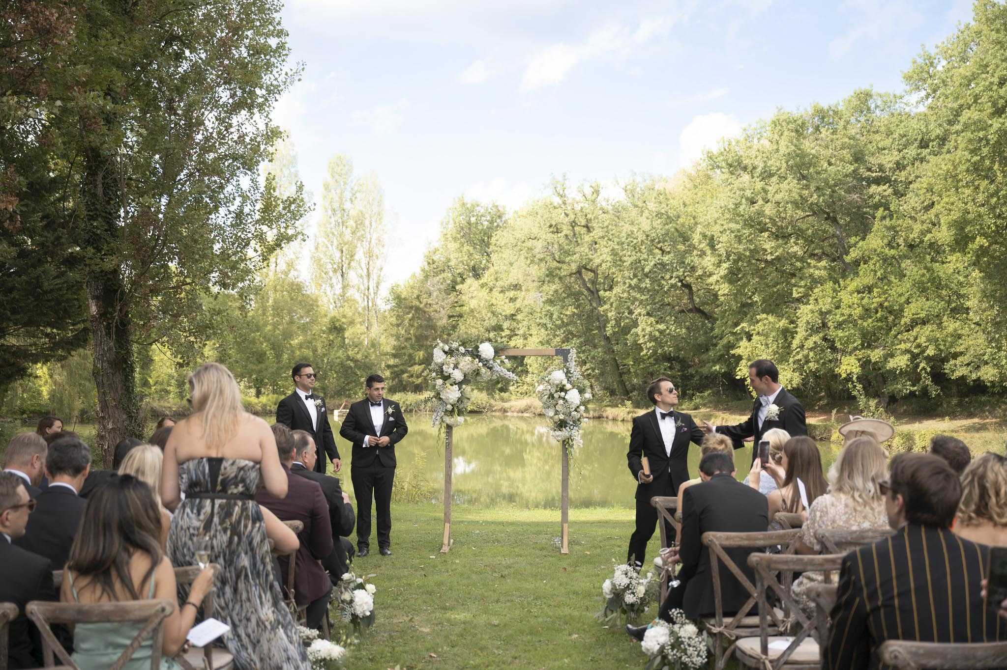 An outdoor wedding ceremony is being set up or is moments before the processional begins, with the groom and three groomsmen — all wearing black tuxedos with bow ties — positioned near the altar and along the aisle. The ceremony takes place on a lawn beside a calm pond, with a wooded backdrop. The altar is a simple wooden square arch adorned with white floral clusters including what appear to be white hydrangeas, roses, and baby's breath, with soft blue-grey accents. Aisle arrangements of white baby's breath and white blooms in low clusters are placed on the ground at the base of guest rows. Guests — approximately 40-50 visible — are seated on rustic wooden cross-back chairs on both sides of the aisle, dressed in formal attire including black suits and patterned dresses. The wide shot captures the full ceremony space from the back of the aisle looking toward the altar and pond, giving a clear view of the layout, decor, and wooded setting.