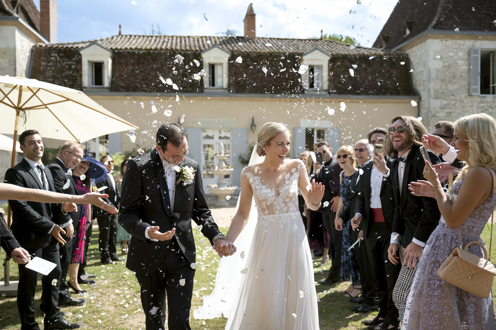 A confetti exit is taking place outdoors in the courtyard of a French manor or chateau, with the couple walking hand-in-hand through two lines of approximately 20 guests who are throwing white flower petals into the air. The bride wears a floor-length white gown with an illusion lace bodice featuring floral appliqué and a flowing skirt, paired with a cathedral-length veil and her hair styled in an updo; the groom wears a dark navy suit with a white boutonniere. Guests are dressed in formal attire including black tuxedos, cocktail dresses in dusty lavender and floral prints, and one woman wearing a navy fascinator hat. The wide-angle shot captures the joy of the moment with both the couple and guests laughing and clapping, with the pale blue-shuttered manor facade and a stone fountain visible in the background.