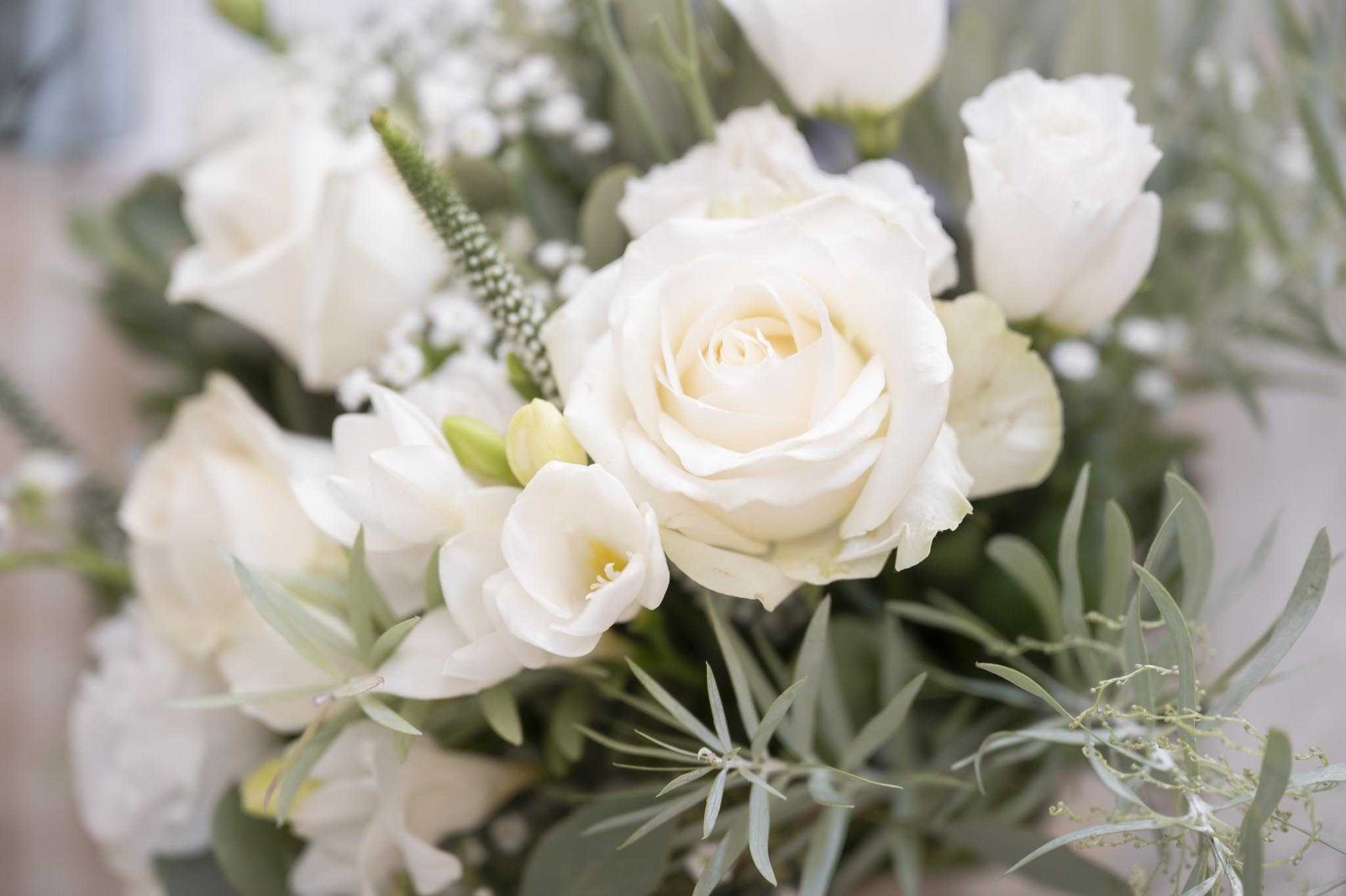 Close-up detail shot of a bridal bouquet composed of ivory roses, white freesias, white tulips, baby's breath, and veronica spikes, with trailing olive branches and wispy greenery throughout. The all-white and ivory floral palette is paired with soft sage-toned foliage, giving the arrangement a classic yet garden-inspired feel. The background is softly blurred, keeping full focus on the central rose bloom. The composition is a tight macro-style detail shot with shallow depth of field.