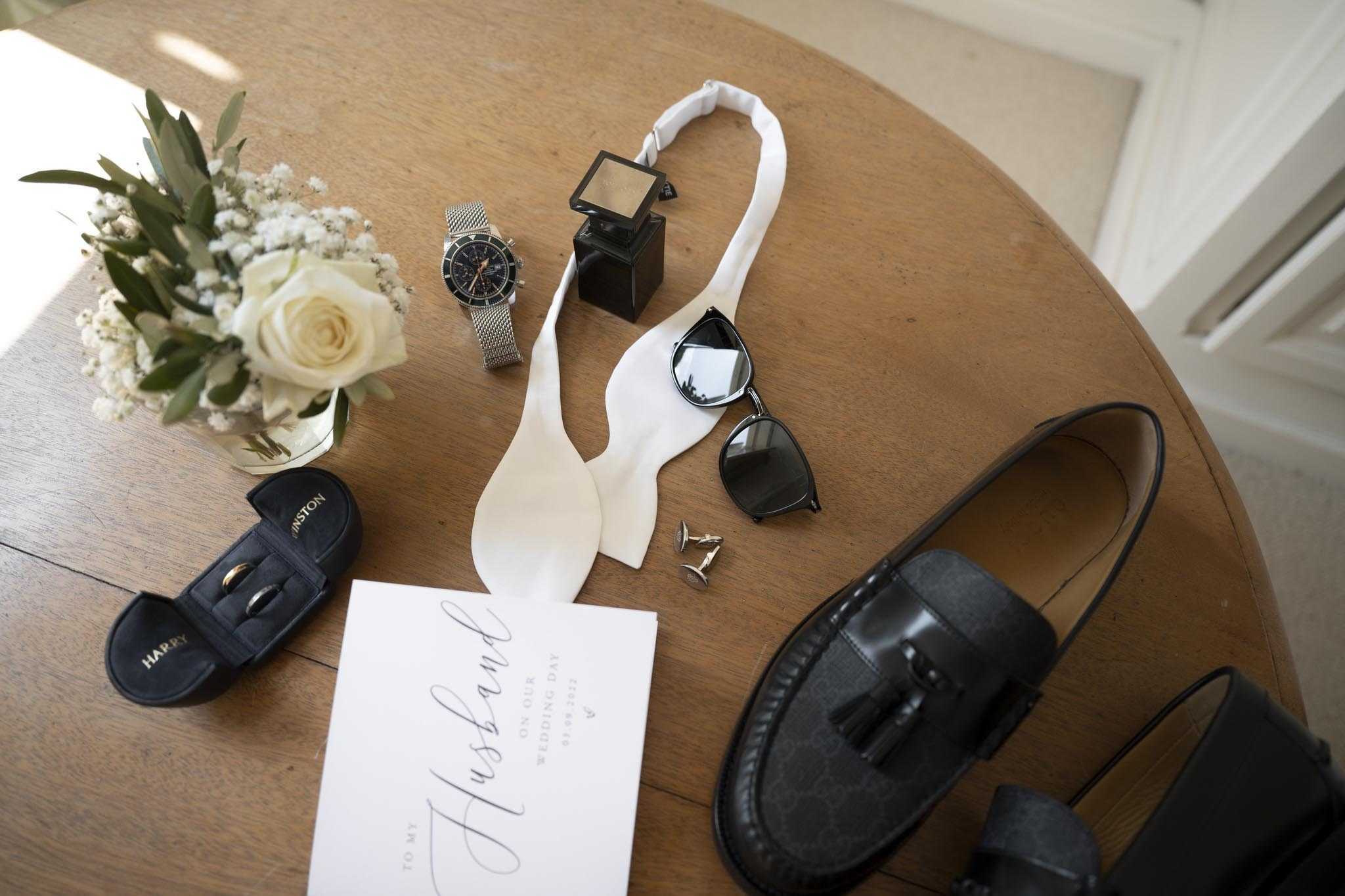 A groom's getting-ready flat lay arranged on a round wooden table, shot from above. The items include a pair of black tassel loafers, a white self-tie bow tie, a silver mesh-strap chronograph watch, a dark square fragrance bottle, black-framed sunglasses, a pair of silver cufflinks, a personalised black ring box labeled 'Harry' containing a gold wedding band, a white calligraphy card reading 'To my Husband on our Wedding Day,' and a small posy of ivory roses, gypsophila, and eucalyptus. The overall styling palette is black, white, and silver with a classic, clean aesthetic.