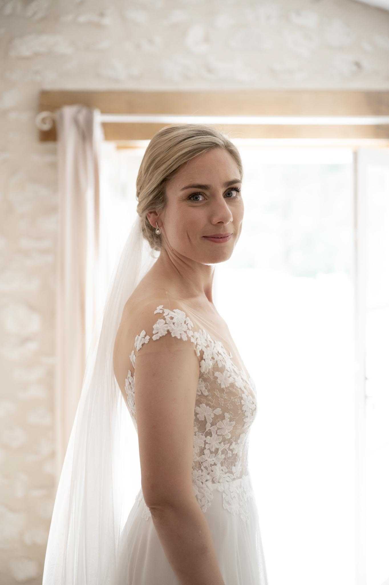 A bridal portrait taken indoors, likely in a getting-ready suite, with the bride positioned in three-quarter view and glancing back toward the camera. She wears an ivory lace wedding gown with an illusion bodice featuring floral lace appliqués extending over the shoulders, paired with a plain-edge cathedral-length veil. Her blonde hair is styled in a low chignon, and she wears small pearl stud earrings with natural, polished makeup. The room features pale floral-patterned wallpaper and blush-toned curtains, with soft natural backlight from a large window creating a bright, airy fill. The shot is a close-up portrait with shallow depth of field.