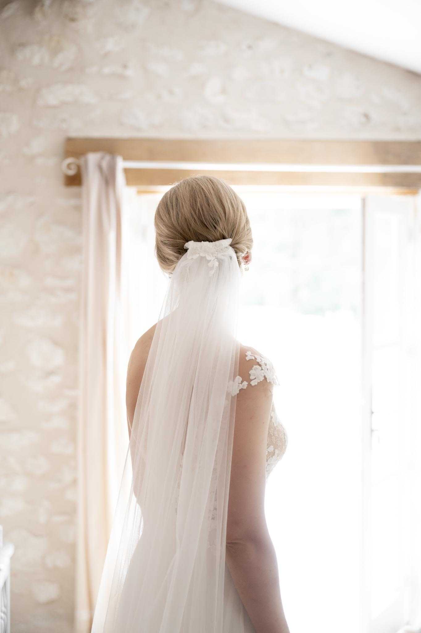 A getting-ready portrait of a bride shot from behind, standing in front of a bright window in an indoor room with pale stone or textured plaster walls and warm wooden window framing. The bride wears an ivory lace wedding dress with illusion cap sleeves featuring floral appliqué detail, and a long, plain-edge tulle veil attached low at the nape of her neck with a small floral hair comb. Her blonde hair is styled in a smooth, low chignon. The composition is a close-up portrait emphasizing the back detail of the dress, veil, and hairstyle, with natural backlight creating a soft, washed-out glow through the window.