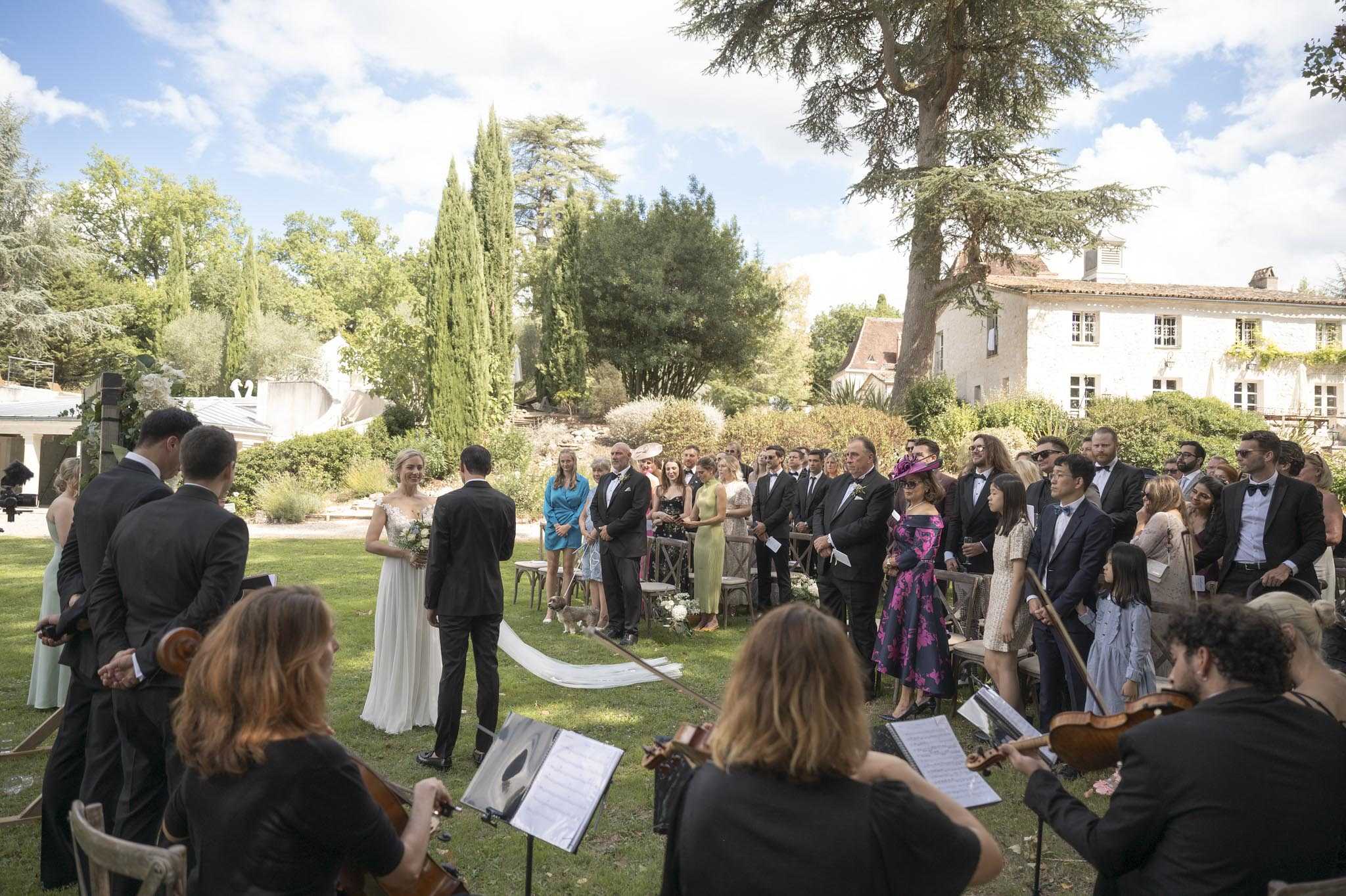 An outdoor wedding ceremony taking place on a lawn in front of a traditional French stone manor house, with approximately 50 guests standing and seated in wooden cross-back chairs arranged in rows on either side of the aisle. The bride wears a white lace cap-sleeve gown with a cathedral-length train and carries a white and greenery bouquet, standing facing the groom who is dressed in a black tuxedo. In the foreground, a string quartet — two visible musicians playing violin and cello, dressed in black — perform with sheet music on stands, shot from behind. Guests are dressed in formal attire including dark suits and tuxedos, with several women in colorful occasion dresses including a bright fuchsia floral midi dress and a teal-blue dress. Bridesmaids in sage green dresses are visible to the left side. The setting is a classic French country estate garden with cypress trees framing the scene, and the ceremony has a classic, formal style.