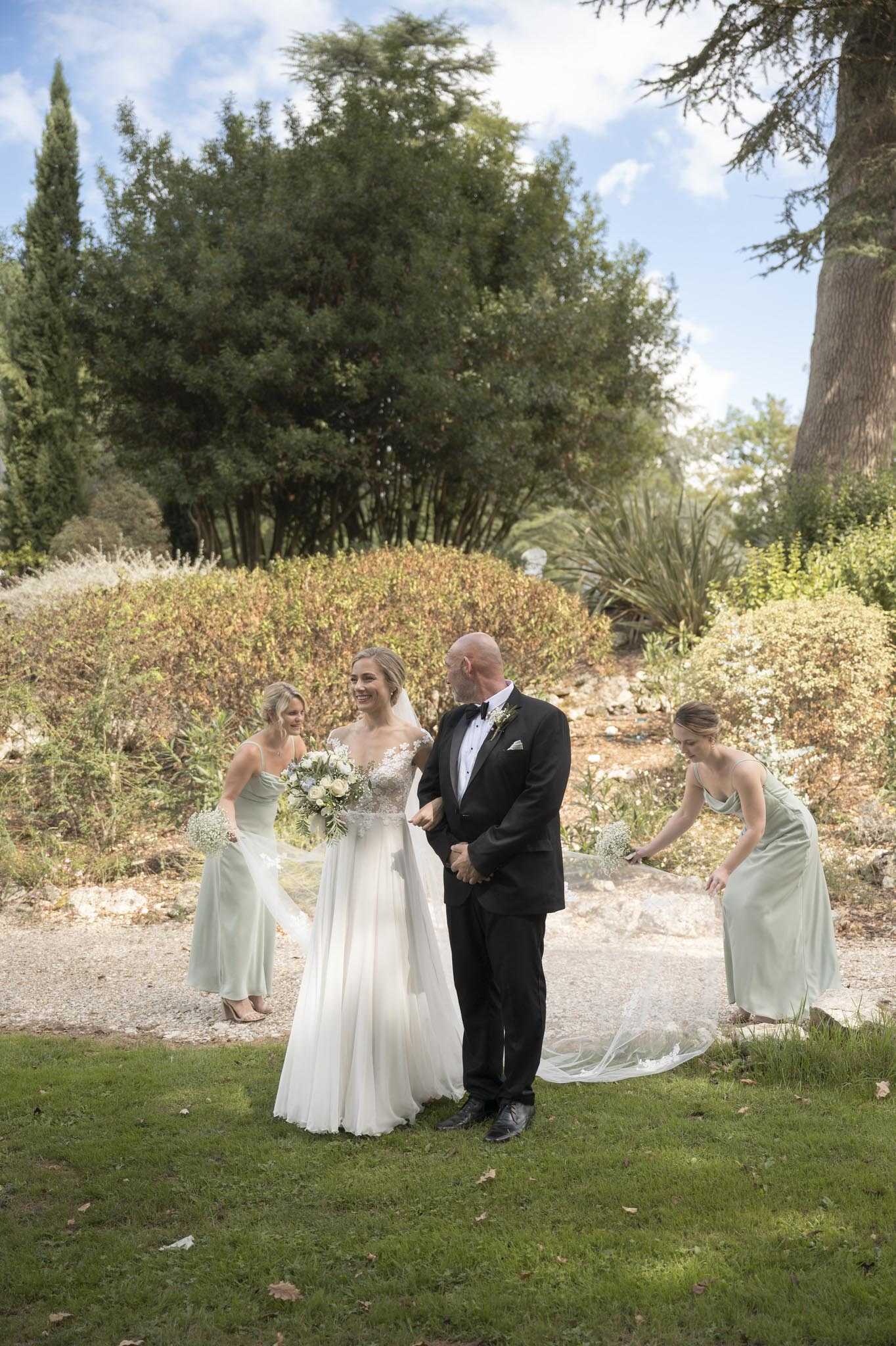 A bride is being escorted by an older man in a black tuxedo with bow tie — likely her father — along a gravel garden path, in what appears to be a pre-ceremony processional moment outdoors. The bride wears a white A-line gown with a lace appliqué bodice and a long flowing tulle skirt and veil, and carries a bouquet of white blooms and greenery including what appears to be baby's breath and eucalyptus. Two bridesmaids in sage green satin slip-style dresses flank the couple — one on the left holds a small bouquet of baby's breath, while the one on the right bends down to arrange the bride's veil and train. The shot is a medium wide portrait taken at garden level, capturing all four figures against a backdrop of sculpted hedges and cypress trees typical of a French estate garden.