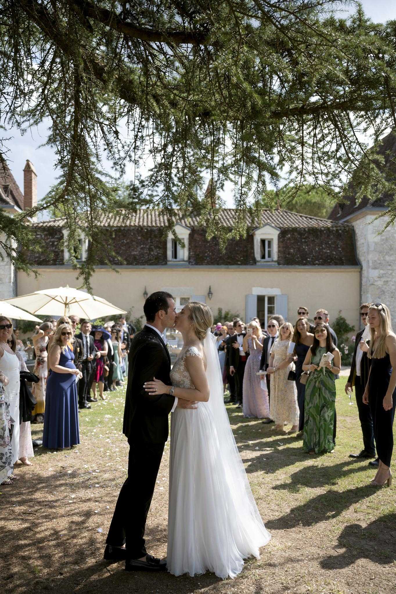 The bride and groom share a kiss outdoors on the grounds of a French chateau during what appears to be a post-ceremony celebration, with approximately 30 guests lined up on either side forming a loose aisle. The bride wears a white tulle A-line gown with a lace-embellished bodice and a cathedral-length veil, while the groom is dressed in a dark navy suit. Guests are dressed in smart-casual to formal attire, with several women in floor-length gowns in shades of cobalt blue, green floral print, and champagne. The setting is a classic French country estate with terracotta-tiled rooflines, pale stone walls, and blue-grey shuttered windows visible in the background, with a cream market umbrella set up to one side. The shot is taken from a medium-wide angle framing the couple centrally beneath the overhanging branches of a large conifer tree.