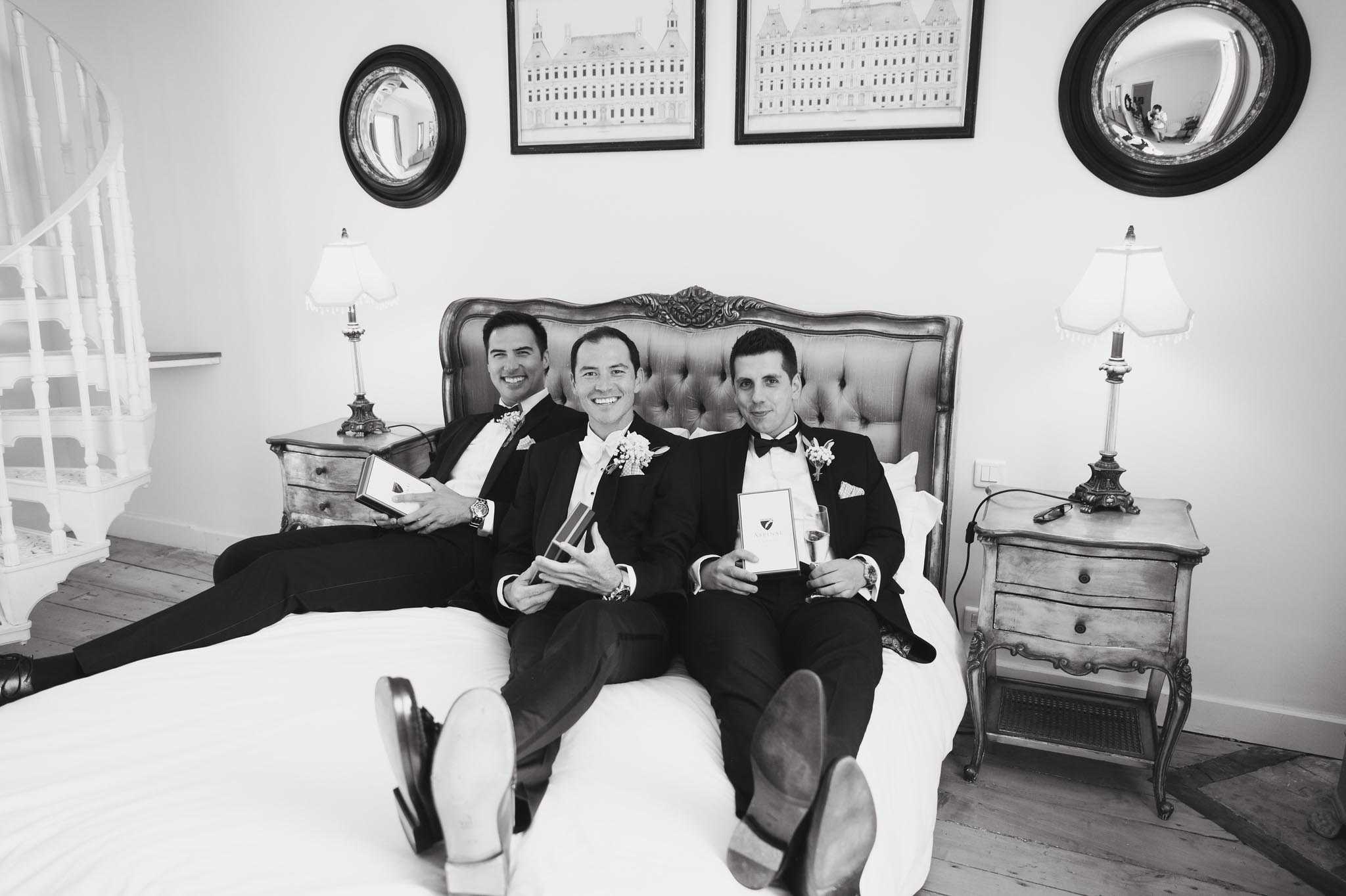 Black-and-white getting-ready shot of three men — likely the groom and two groomsmen — lounging casually on a tufted upholstered bed in a chateau-style bedroom. All three wear black tuxedos with bow ties, white dress shirts, and boutonnières featuring small white floral clusters; two of the men hold gift boxes and one holds a champagne flute. The room features French provincial-style bedside tables with traditional table lamps, two round convex mirrors on the wall, and two framed architectural prints depicting what appears to be a large historic building; a white spiral staircase is partially visible on the left. The image is shot from a low angle at the foot of the bed, showing the soles of their shoes in the foreground, giving the composition a playful, candid feel. High contrast black-and-white tones give the image a sharp, graphic quality.