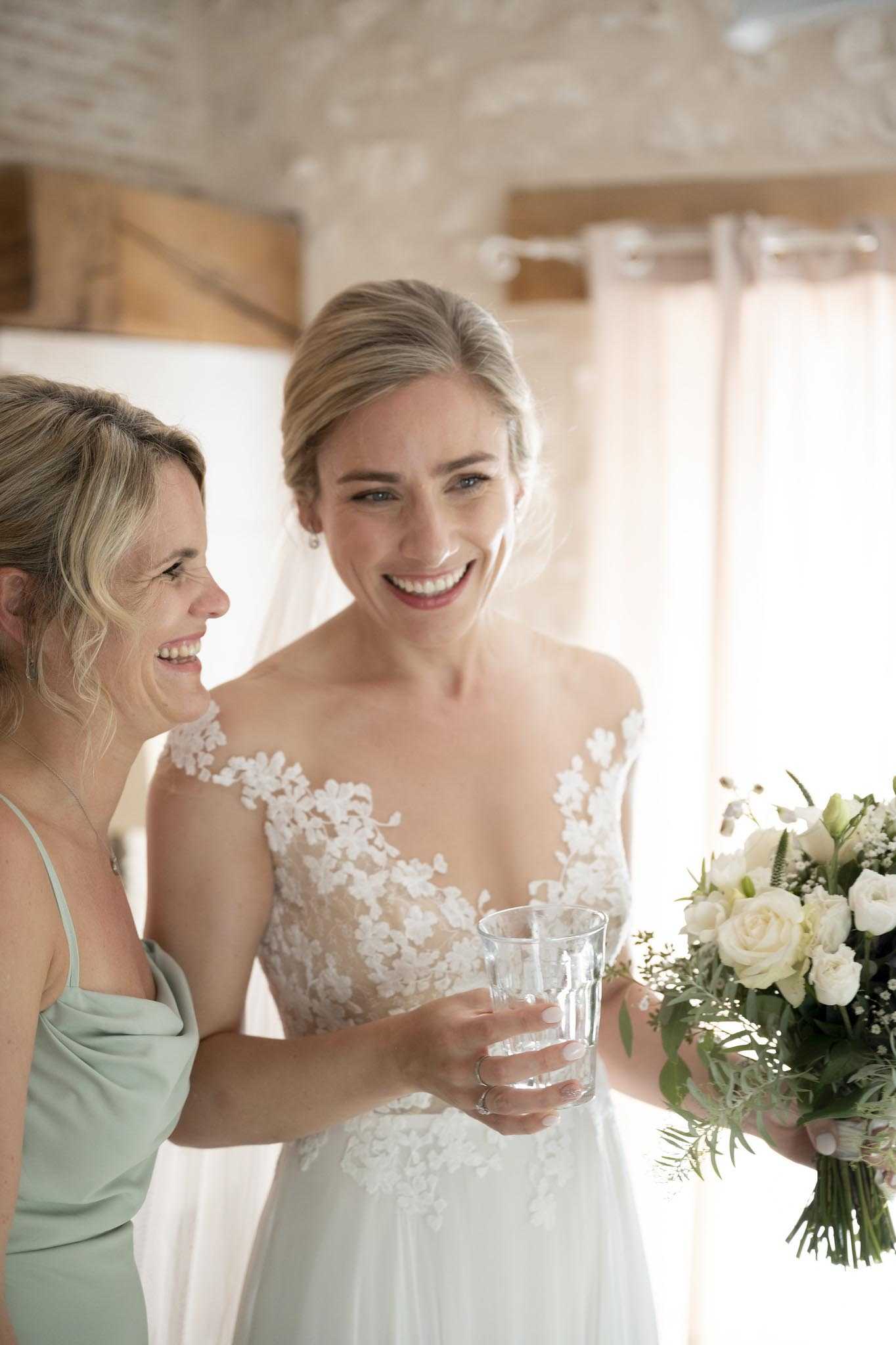 A candid getting-ready moment showing the bride and one bridesmaid laughing together indoors, likely in a rustic venue with exposed stone walls and wooden beams visible in the background. The bride wears a white gown with an illusion lace bodice featuring floral appliqué detailing over the neckline and shoulders, and holds a glass of water in one hand and her bouquet in the other; the bouquet consists of ivory roses, white lisianthus, gypsophila, and green foliage. The bridesmaid wears a sage green cowl-neck slip-style dress. The shot is a medium portrait with soft natural backlight from a sheer-curtained window, giving the image a bright, airy feel.