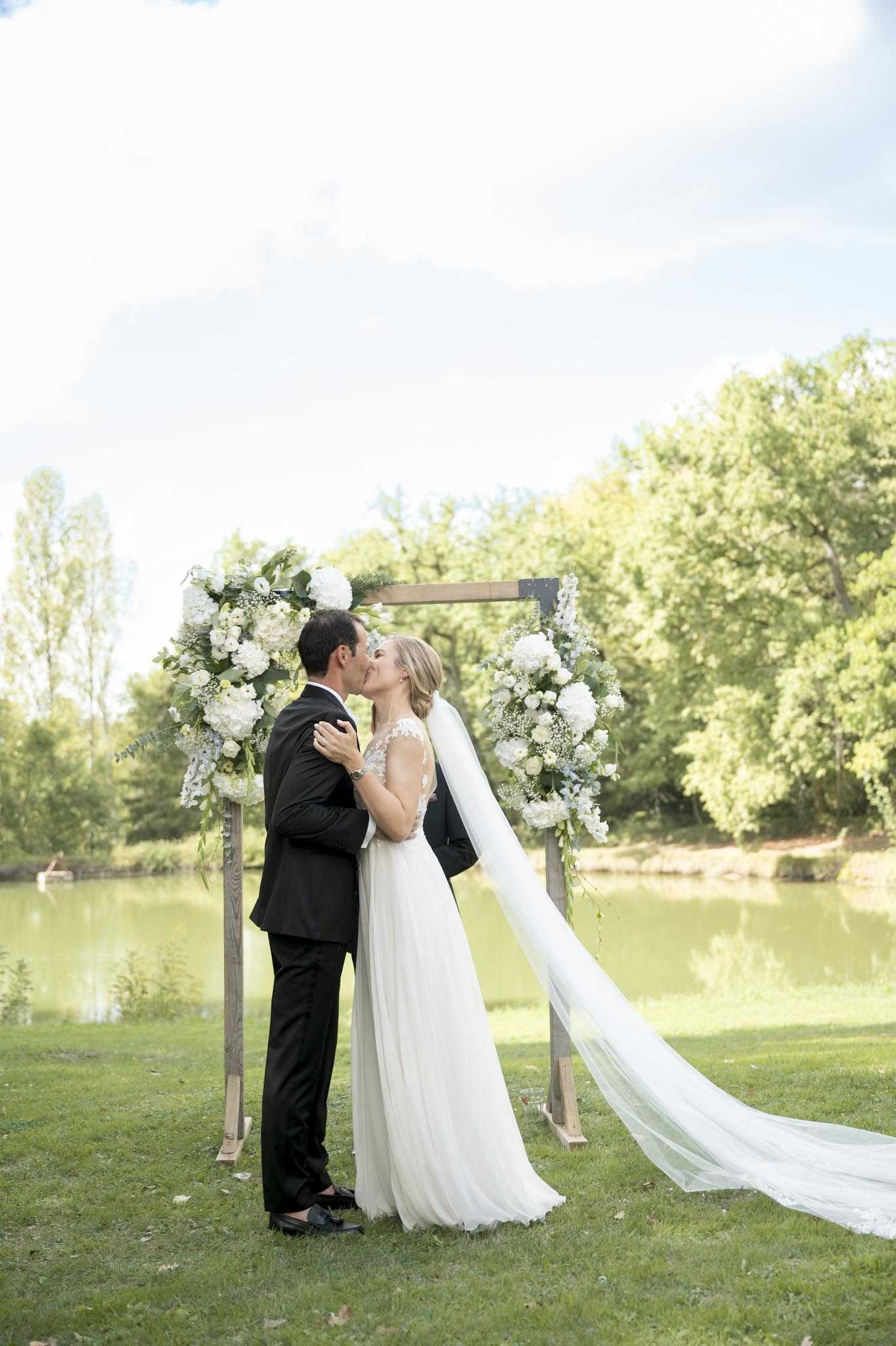 A couple shares their first kiss during an outdoor ceremony on a lawn beside a calm pond. The groom wears a black suit and the bride wears a white A-line gown with a lace bodice and an extra-long cathedral veil that trails across the grass, caught slightly by a breeze. They stand beneath a square wooden arch decorated with large clusters of white hydrangeas, white ranunculus, baby's breath, and lush green foliage on each lower corner. The overall floral and decor palette is white and green with a clean, classic style. The shot is a medium full-length portrait framing both figures against the arch and waterside backdrop.