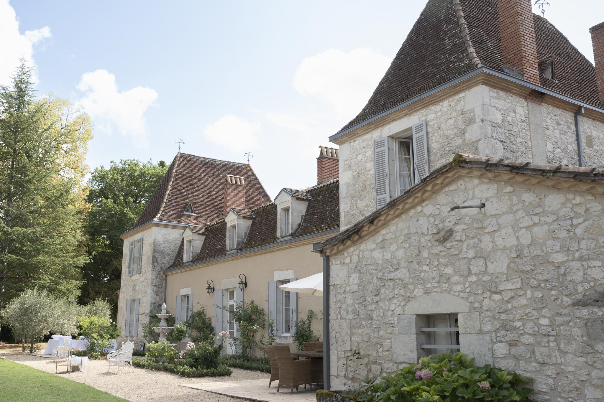 Outdoor wide shot of a French chateau complex featuring multiple stone and rendered limestone buildings with steep terracotta-tiled roofs, conical towers, dormer windows, and grey-painted shutters. The grounds include a gravel terrace with rattan lounge chairs under a large white parasol, wrought-iron garden furniture, a stone pedestal fountain, and climbing roses along the facade. In the background to the left, white-draped chairs or table linens are visible, suggesting wedding setup in progress. The overall styling of the venue decor is classic French country, with soft pink climbing roses and hydrangeas adding color accents to the exterior. Potential venue feature image.