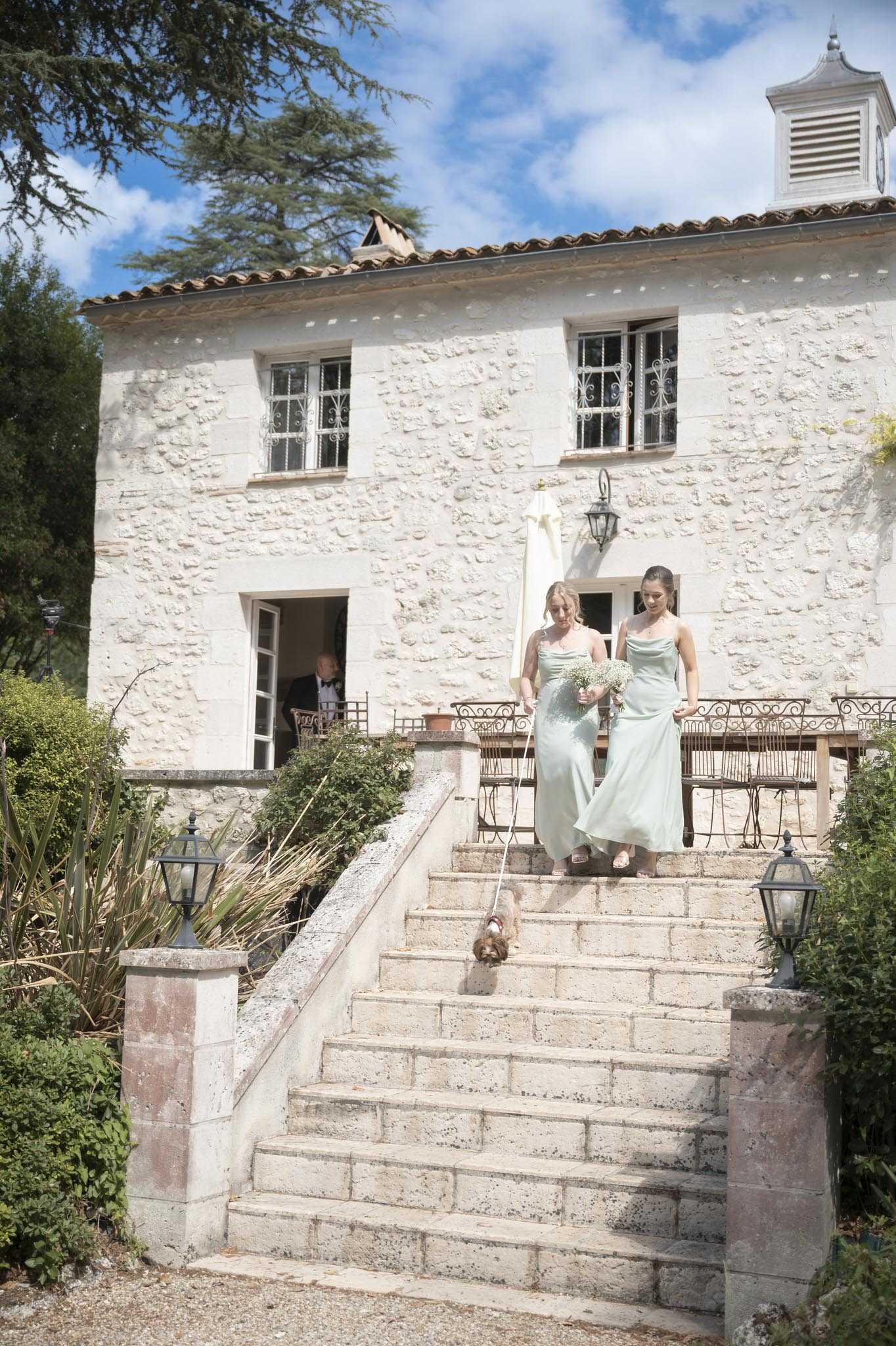 Two bridesmaids in matching sage green satin cowl-neck floor-length dresses descend a wide stone staircase at a French country property, one carrying a small bouquet of white baby's breath and walking a small dog on a lead. A man in a black tuxedo is visible in the background through an open doorway at the top of the terrace. The venue is a two-storey limestone building with terracotta roof tiles, ornate wrought-iron window grilles, a small cupola, and stone pillar lanterns flanking the staircase, with wrought-iron bistro furniture visible on the upper terrace. The shot is a medium wide-angle taken from the base of the steps, showing the full facade of the building.