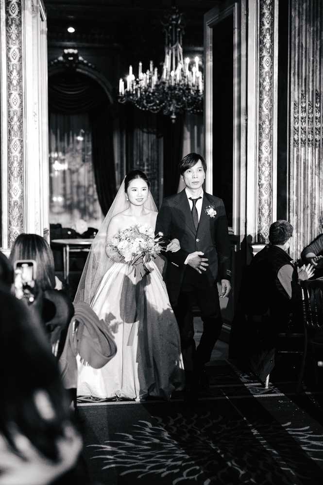 Black-and-white image of a bride walking down the aisle escorted by a man in a dark suit with a boutonniere, likely her father, during an indoor ceremony. The bride wears a strapless ball gown with a long veil and carries a full, loosely arranged bouquet with mixed blooms and trailing elements. The setting is a grand ballroom or event hall with ornate architectural details including tall mirrored pilasters, heavy draped curtains, and a crystal chandelier visible in the background, conveying a classic formal aesthetic. The shot is taken from a medium distance at guest level, with seated and standing guests visible in the foreground capturing the moment on their phones, and the contrast between the bright white of the bride's gown and the dark surroundings draws the eye to the couple mid-processional.