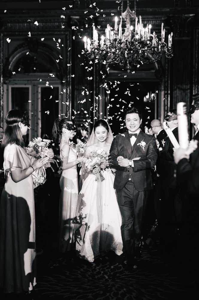 Black-and-white image of a couple making their recessional walk through a guest-lined aisle inside an ornate ballroom or grand hall, with white confetti or paper butterflies filling the air around them. The bride wears a strapless ballgown with a long veil and carries a full, loose bouquet, while the groom wears a dark suit with a bow tie and boutonniere. Guests on both sides — including bridesmaids in floor-length gowns holding bouquets — clap and cheer, with some holding white glow sticks or cannons. The interior features high ornate ceilings, arched paneled walls, and a large lit candelabra chandelier overhead, suggesting a classic Haussmann-era Parisian venue. The image is a medium portrait shot with high contrast black-and-white tones, capturing motion and celebration.