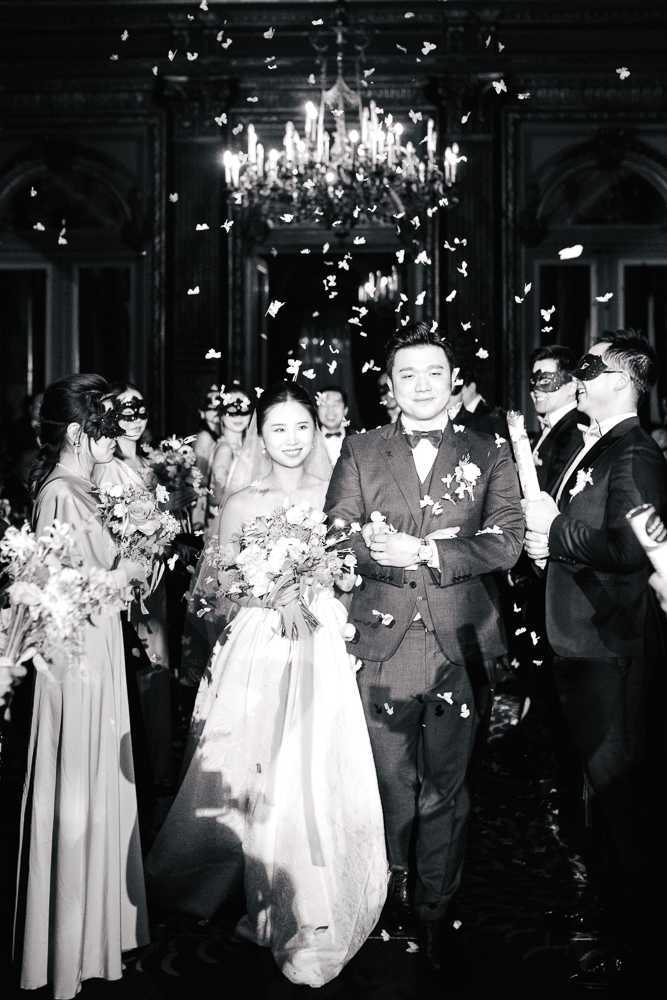 This black-and-white image captures the couple's grand exit through an indoor reception hall, with small butterfly-shaped confetti raining down around them. The bride wears a strapless ballgown and carries a full, loose bouquet, while the groom is dressed in a three-piece suit with a bow tie and boutonnière. Guests lining both sides of the exit path wear masquerade-style black eye masks, giving the event a masquerade ball theme; bridesmaids in floor-length gowns hold bouquets on the left side, while suited male guests hold glow sticks on the right. The ornate interior features dark wood paneling, arched doorways, and a chandelier in the background, suggesting a classic ballroom or château setting, with high contrast between the bright confetti and chandelier light against the dark architectural backdrop.