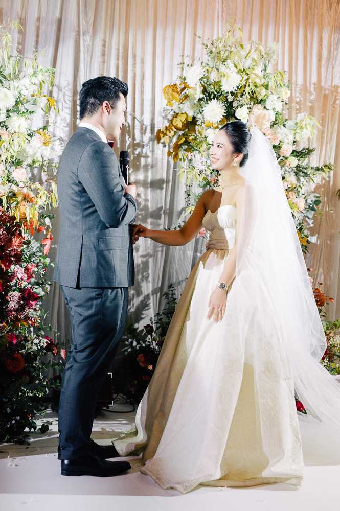 The couple stands face-to-face during an indoor ceremony vow exchange, with the groom in a charcoal grey suit holding a microphone while reading his vows, and the bride smiling up at him while holding his hand. The bride wears a strapless white ball gown with a champagne-gold sash at the waist and a long cathedral veil, with her hair pulled back. They are positioned in front of a large floral installation featuring ivory dahlias, peach roses, burnt orange blooms, deep burgundy florals, and autumn-toned foliage arranged against draped blush and ivory fabric panels. The floral arrangement extends on both sides, creating a rich autumnal color palette of terracotta, rust, deep red, and gold tones. This is a medium portrait shot capturing both figures in full from a slight side angle.