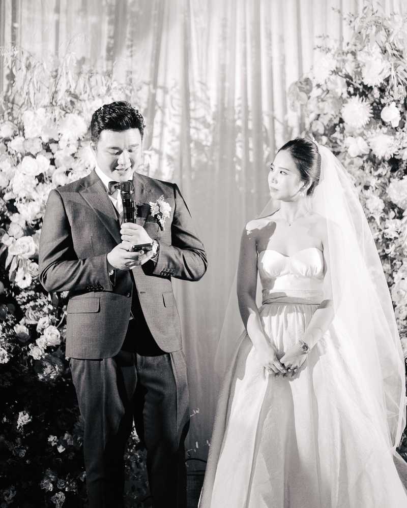 This black-and-white portrait captures the couple during what appears to be the ceremony vow exchange or speech moment, with the groom holding a microphone and reading from his phone while the bride looks at him with an emotional expression. The groom wears a dark suit with a bow tie and a floral boutonniere, while the bride wears a strapless ballgown with a structured bodice and a long cathedral veil, her hair pulled back. They stand in front of a draped curtain backdrop flanked by large, lush floral arrangements featuring dahlias, roses, and leafy branches in light tones against deep foliage. The image is a mid-shot with strong contrast, the couple well-lit against the softer background.