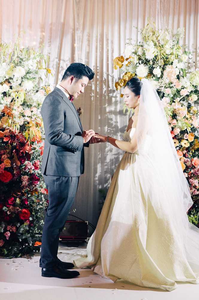 The couple is exchanging rings during an indoor wedding ceremony, with the groom placing the ring on the bride's finger. The groom wears a charcoal grey suit with a burgundy bow tie, while the bride wears a strapless ivory ball gown with a champagne-toned sash at the waist and a long cathedral veil. The ceremony backdrop features floor-to-ceiling floral installations in a rich, autumnal palette of deep red, burnt orange, coral, blush, mustard yellow, and white blooms with lush greenery, flanking a sheer draped curtain. The composition is a full-length portrait shot capturing both figures against the floral backdrop.
