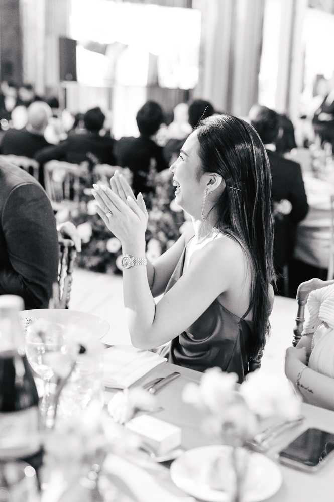 A black-and-white image captured during a wedding reception, showing a female guest seated at a dining table clapping and laughing, caught in a candid moment of enjoyment — likely during speeches or a performance. She is wearing a sleeveless satin slip-style dress, a bracelet watch, dangling earrings, and a necklace, with long straight hair worn loose. The foreground shows a place setting with glassware, a wine bottle, a favor box, and soft floral arrangements rendered in light mid-tones. The background reveals a large indoor ballroom-style reception venue filled with other seated guests in formal attire, with tall windows and warm ambient lighting creating bright highlights in the high-contrast black-and-white tones. The shot is taken from a slightly elevated angle at a three-quarter perspective, giving it a journalistic, candid quality.