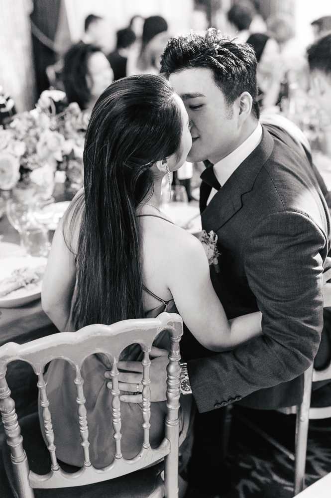 A black-and-white close-up portrait of a couple sharing a kiss at a wedding reception, seated at a decorated dining table. The woman wears a strappy dress and has long dark hair worn down, while the man wears a fitted suit jacket with a dark tie and a small floral boutonniere. They are seated on ornate spindle-back Napoléon-style chairs, with floral centerpieces, glassware, and plates visible along the table behind them. Other guests are visible in the soft-focus background, suggesting a well-attended indoor reception.