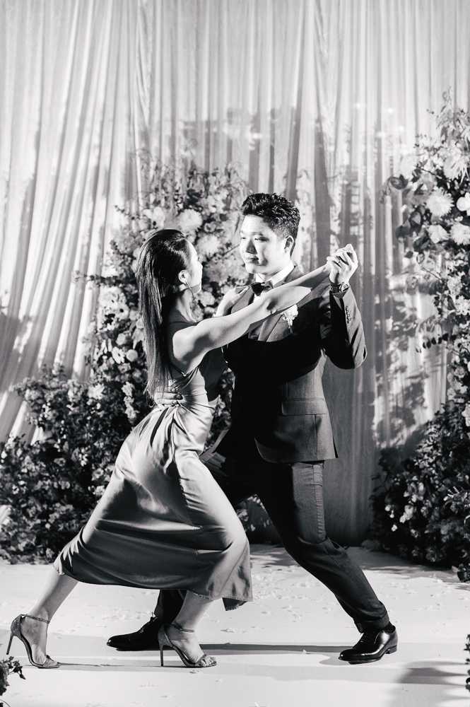 This black-and-white image captures a couple performing a choreographed first dance at an indoor wedding reception. The man wears a dark fitted suit with a bow tie and boutonniere, while the woman wears a satin midi dress with a wrap-style waist and strappy heeled sandals; both are in a wide, low dance stance suggesting a tango or Latin-style routine. The backdrop consists of floor-length white draping flanked by large floral arrangements featuring full blooms and lush greenery, visible in soft mid-tones against the bright curtain. The composition is a full-length portrait shot with strong contrast, emphasizing the dynamic movement and energy of the dance.