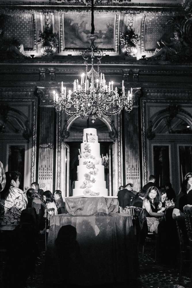 A black-and-white image of a tall five-tier wedding cake displayed on a draped table during an indoor reception. The cake is decorated with large floral appliqués cascading down its tiers, appearing light against the white fondant. The setting is a grand ballroom with ornate architectural details including gilded moldings, large arched doorways, patterned wall panels, painted ceiling panels, and a large candelabra-style chandelier with lit candles hanging directly above the cake. Dozens of guests in formal attire are seated at tables surrounding the cake, visible in the mid and background. The image has deep contrast with dark shadows in the room periphery and bright highlights on the cake and chandelier, giving the scene a classic, high-contrast look. Wide shot composition with the cake as the central focal point.