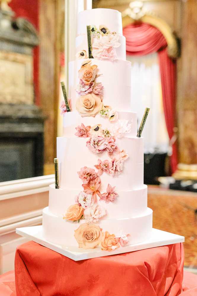 Close-up detail shot of a six-tier white fondant wedding cake displayed on a white square base atop a coral-red damask tablecloth. The cake is decorated with a cascading arrangement of fresh flowers in peach roses, blush and dusty pink peonies, mauve tulips, and white anemones with dark centers, trailing diagonally from the top tier to the base. Small dark green decorative rods are inserted between tiers as accents. The setting is an indoor ballroom or reception room with ornate gold molding, a fireplace, and red draped curtains visible in the soft-focus background, suggesting a classic or grand historic venue interior.