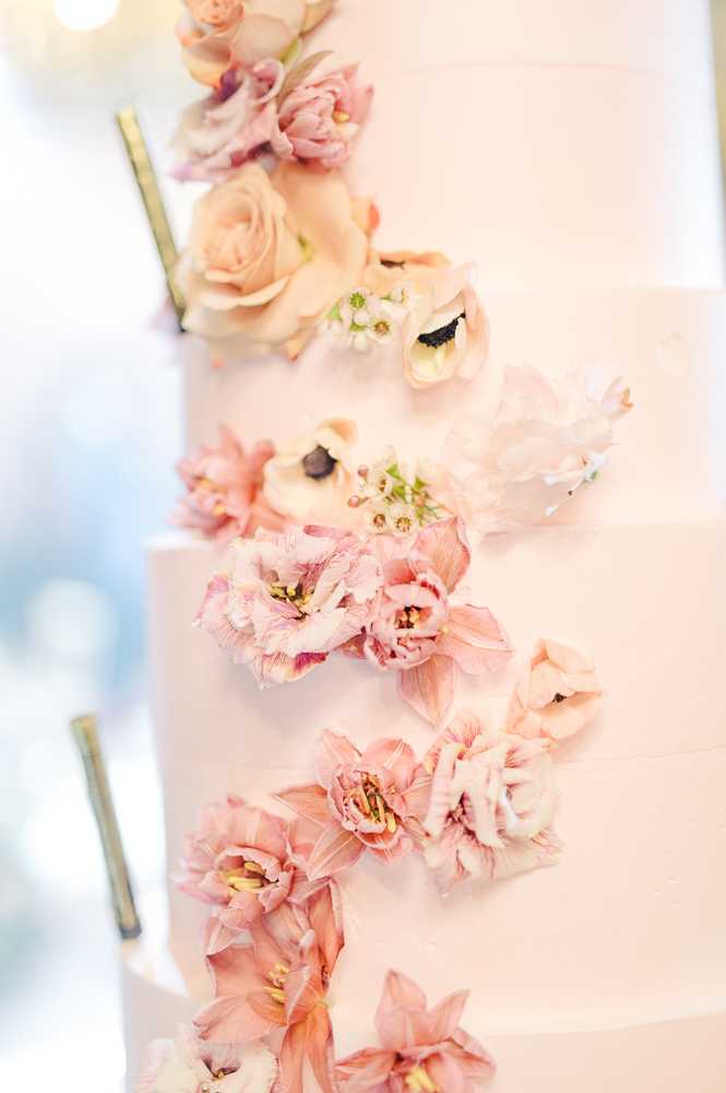 Close-up detail shot of a multi-tiered white wedding cake decorated with a cascading arrangement of fresh flowers along one side. The floral cascade includes blush and peach garden roses, mauve-pink amaryllis blooms, white anemones with dark centers, small white wax flowers, and delicate blush parrot tulips. The cake itself has a smooth white finish, and the flowers are arranged in a diagonal trailing pattern from the top tier downward. The background is softly blurred with light bokeh, keeping the focus entirely on the cake and florals.