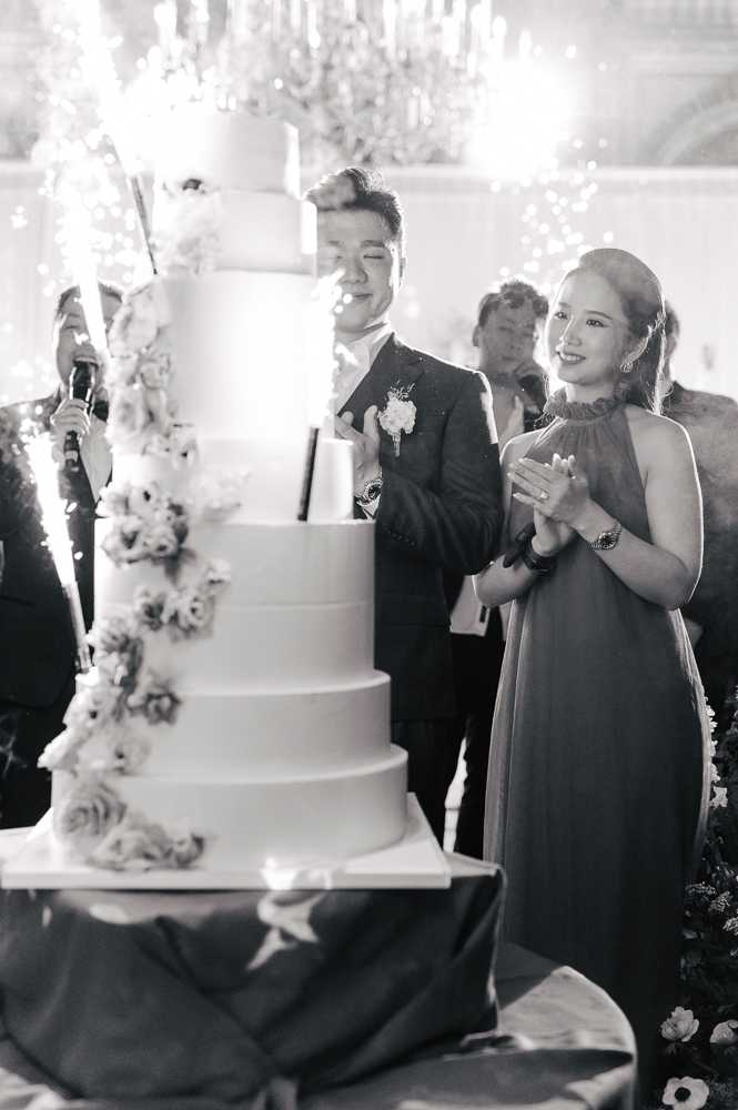 This black-and-white image captures a cake cutting moment at an indoor wedding reception, set in what appears to be a formal ballroom with an ornate crystal chandelier visible in the background. A tall, six-tier wedding cake decorated with cascading floral arrangements stands prominently in the foreground, with sparklers shooting light from its top tiers. The groom, wearing a dark suit with a floral boutonniere, stands beside the bride, who is dressed in a halter-neck gown and is clapping her hands with a wide smile; several guests are partially visible behind them. The overall styling is classic and formal, with high contrast tones in the image emphasizing the sparkler light and the cake's smooth tiered surface against the bright chandelier backdrop.