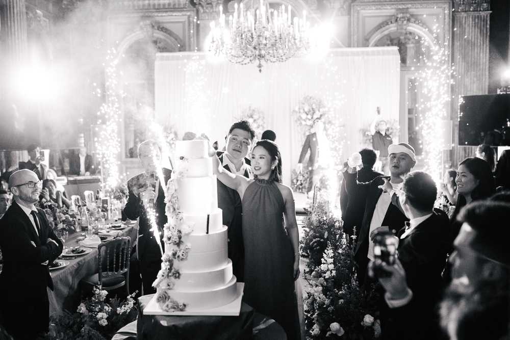 Black-and-white image of a cake cutting moment during an indoor wedding reception in an ornate ballroom featuring a crystal chandelier, tall decorative columns, and draped fabric backdrop. The couple stands before a tall five-tier white wedding cake adorned with floral accents cascading down one side, while sparklers ignite around them creating dramatic light bursts and bright highlights in the frame. The groom is dressed in a dark suit and the bride wears a halter-neck gown, surrounded by a group of attendants in formal attire including white bow ties, as well as seated guests at candlelit dinner tables visible to the left. The wide-angle shot captures the high energy of the moment, with guests photographing on phones and the overall decor suggesting a formal, black-tie classic wedding aesthetic with lush floral arrangements lining the foreground.
