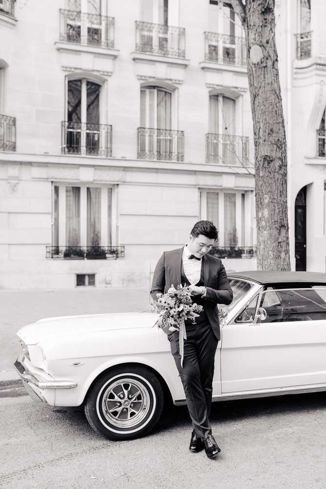 A black-and-white portrait of a groom standing outdoors on a Parisian street, leaning against a vintage Ford Mustang convertible with a dark soft top and white-wall tires. The groom wears a dark slim-fit suit with a bow tie and a floral or patterned dress shirt, and holds a loose, trailing bridal bouquet with long ribbon streamers while looking downward. Behind him, a classic Haussmann-style building with tall arched windows and wrought-iron balconies forms the backdrop. The image has bright, high-key tones with soft contrast, and is composed as a full-length portrait shot from street level.