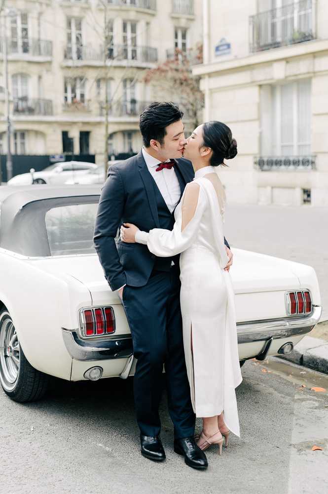 A couple shares a kiss during an outdoor portrait session on a Parisian street, leaning against a vintage cream Ford Mustang. The bride wears a minimalist white long-sleeve midi dress with a sheer back panel and a front slit, paired with nude heeled sandals and her hair styled in a low updo. The groom wears a navy three-piece suit with a white dress shirt and a deep red bow tie, finished with black oxford shoes. The setting features classic Haussmann-style building facades in the background, giving the shoot a modern, city-chic aesthetic. The image is a full-length couple portrait with a soft, light editorial tone.