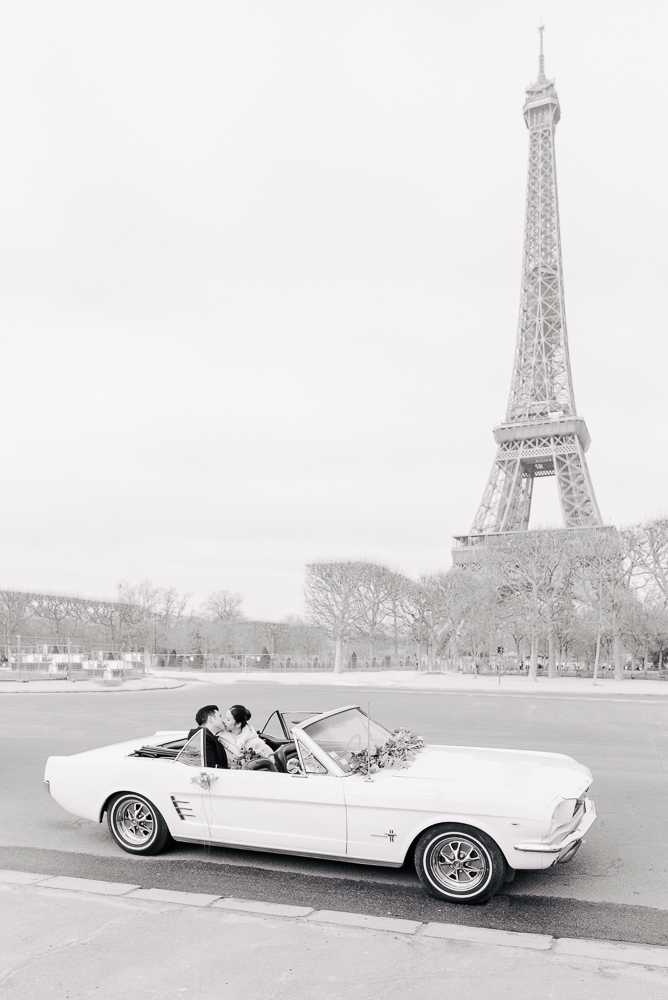 This black-and-white image shows a couple kissing in a vintage convertible Ford Mustang parked on a street in Paris, with the Eiffel Tower prominent in the background. A bridal bouquet is visible resting on the dashboard or door of the open-top car. The image is rendered in soft, high-key tones with bright whites and gentle mid-range contrast. The composition is a wide portrait-oriented shot taken from street level, capturing the full length of the classic white Mustang with the tower rising behind it, framed by bare-branched trees lining the road.