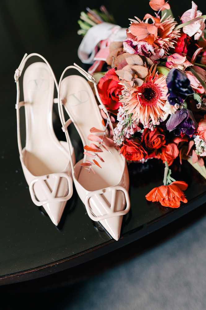 A close-up detail shot of a pair of nude patent leather Valentino VLogo slingback heels placed on a dark round surface alongside a bridal bouquet. The bouquet features a bold, jewel-toned palette of red roses, coral gerbera daisies, deep purple anemones, pink alstroemeria, and terracotta dried botanicals, with a few loose petals scattered across the surface. The shoes display the signature Valentino VLogo cut-out hardware at the toe in matching nude patent leather. The dark background and surface provide high contrast against the light-colored shoes and vibrant florals.