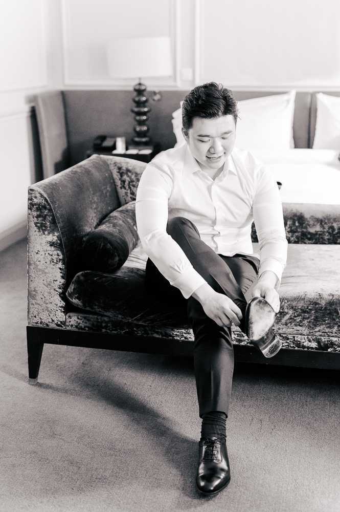 Black-and-white getting-ready portrait of a groom seated on a velvet upholstered sofa in what appears to be a hotel suite, putting on his dress shoes. He is wearing a white dress shirt and dark trousers, with one polished oxford shoe already on and the other being fastened. The room features a sculptural table lamp, side table, and a bed partially visible in the background, rendered in soft mid-tones with gentle contrast. The shot is a full-length portrait with a relaxed, candid quality.