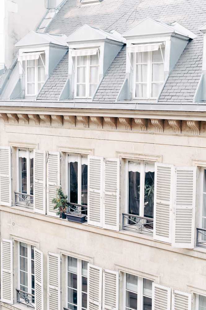 This image shows the exterior facade of a classic Haussmann-style Parisian building, featuring cream limestone walls, white louvered shutters, ornate carved corbels along the roofline cornice, and mansard roof dormers with slate tiles. Small wrought-iron balconies with potted green plants are visible on the middle floor. No people, wedding party, or event activity are present in this image — it appears to be an architectural detail or location shot rather than a wedding photograph. Wide shot, slightly upward angle. Potential venue feature image.