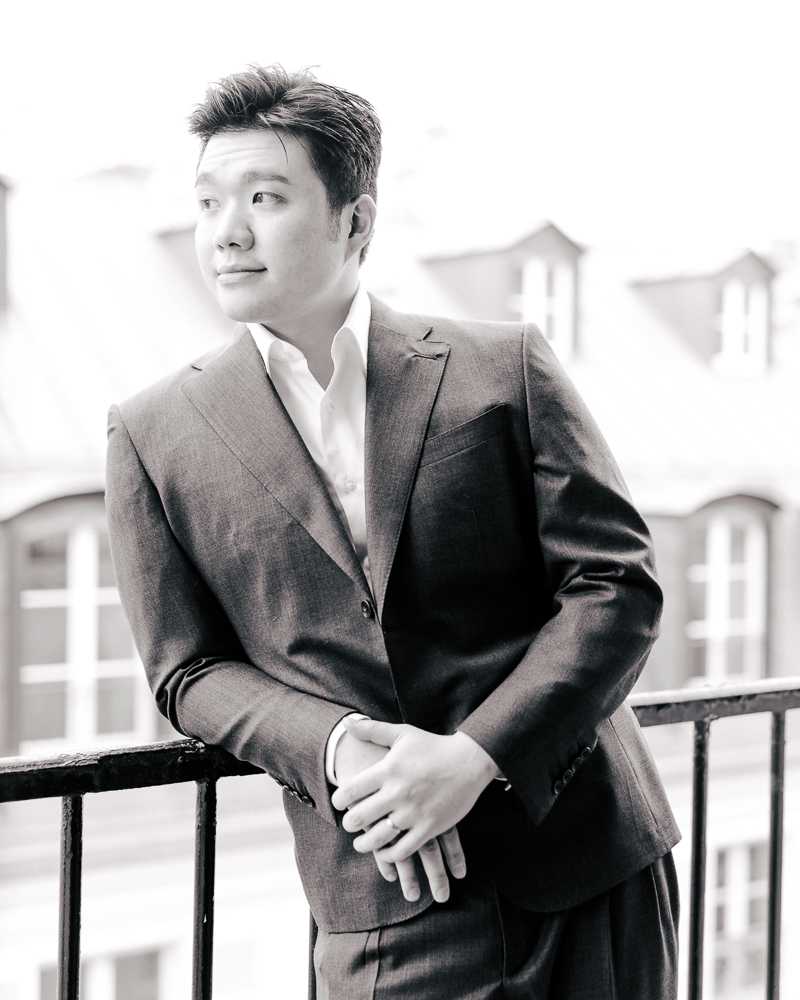 This is a black-and-white portrait of a groom standing on an outdoor balcony or terrace, leaning against a dark metal railing with his hands clasped in front of him and his gaze directed off to the side. He is wearing a dark suit jacket with a white dress shirt, open at the collar with no tie. The image has bright, high-key tones with soft contrast, and Haussmann-style rooftop architecture with mansard roofs and arched windows is visible in the soft-focus background, suggesting a Paris city setting. The composition is a medium portrait shot.