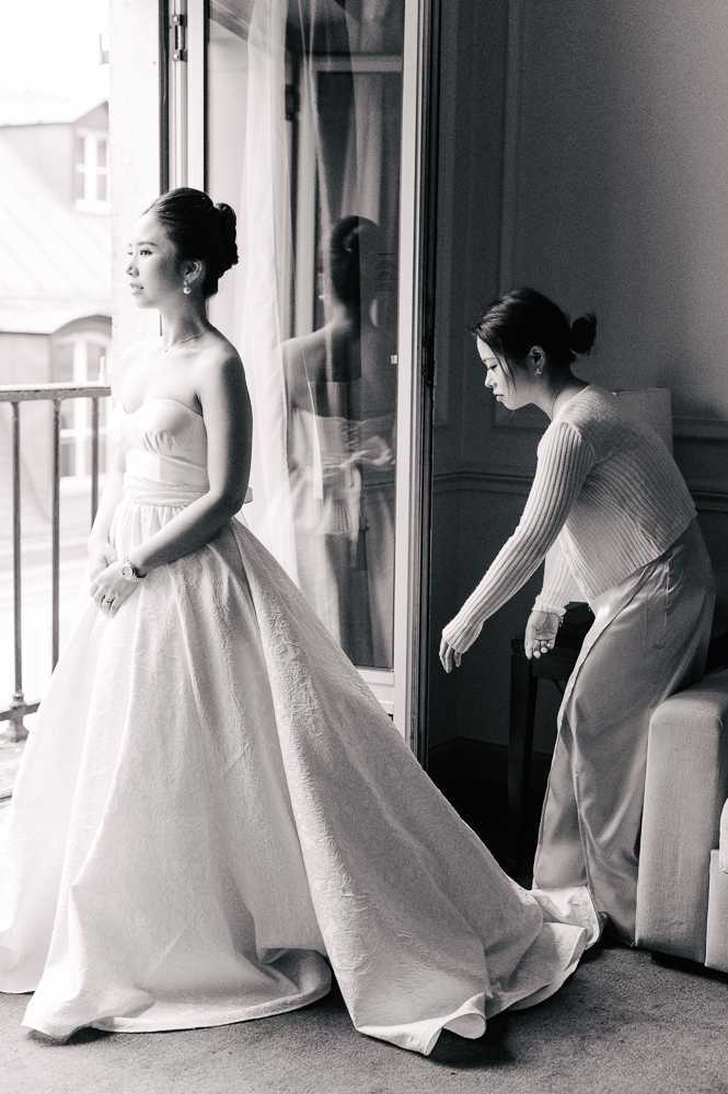 A black-and-white getting-ready shot taken indoors in what appears to be a Parisian hotel room, with tall French doors and a balcony visible in the background. The bride stands in profile near the window, wearing a strapless ballgown with a textured full skirt and a cathedral-length train, her hair styled in a sleek updo with pearl earrings and a delicate necklace. A second woman, dressed in a ribbed long-sleeve top and wide-leg trousers with her hair in a bun, bends down to arrange and spread the train of the dress. The bride's reflection is partially visible in the glass door panel, adding depth to the composition, which is a medium full-length portrait with strong natural backlight creating high contrast between the bright window and the darker interior tones.