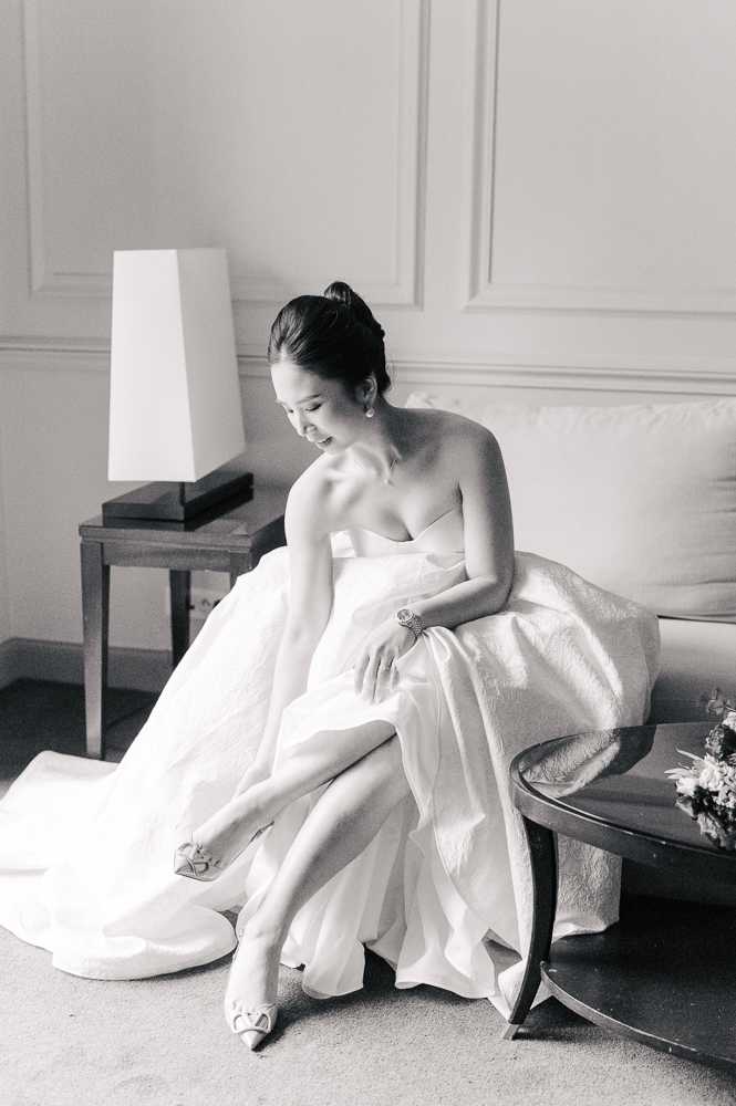 This black-and-white getting-ready portrait shows a bride seated on a sofa in an indoor room with classic paneled walls, adjusting her heeled bridal shoes. She wears a strapless ballgown with a full skirt and a structured sweetheart neckline, paired with pointed-toe heels featuring a bow detail, drop earrings, and a bracelet watch. Her dark hair is pulled up into a sleek high bun, and she is smiling downward. A table lamp and side table are visible to her left, and a small floral arrangement sits on a low table to her right. The image uses soft, bright tones with gentle contrast typical of film-style photography, and is composed as a three-quarter portrait shot.