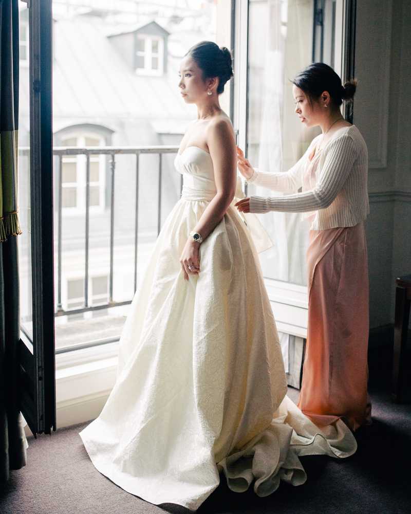 A getting-ready scene shot indoors in what appears to be a Parisian hotel room, with French doors open to a wrought-iron balcony and Haussmann-style rooftops visible in the background. The bride stands in profile wearing a strapless ivory ballgown with a structured bodice and full textured skirt with a train pooling on the floor, accessorized with pearl drop earrings and a dark-faced watch. A second woman in a blush-pink maxi skirt and cream ribbed knit top stands behind the bride, fastening or adjusting the back of the gown. The composition is a full-length portrait shot with soft natural window light illuminating both figures from the front.