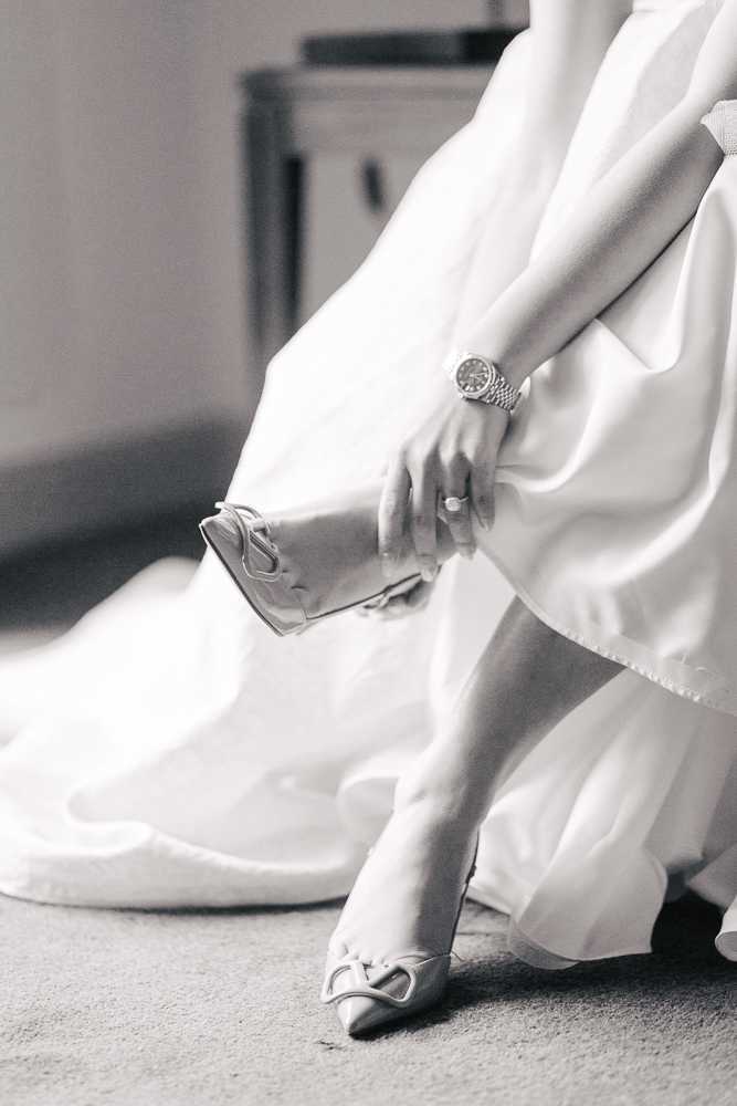 A black-and-white close-up detail shot of a bride getting ready indoors, captured from the waist down. She is seated and wearing a full-skirted white bridal gown with a voluminous skirt, and is in the process of fastening a strappy heeled shoe with a decorative buckle detail at the toe. Her hand is visible wearing a silver bracelet-style watch and an engagement ring. The image uses soft mid-tones with bright highlights on the satin fabric of the dress, creating a clean contrast against the darker background.