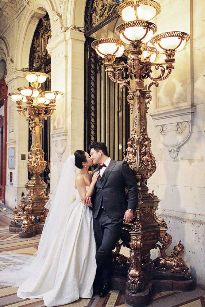 A couple shares a kiss during a wedding portrait session inside an ornate Beaux-Arts building, likely the Palais Garnier or a similar grand Parisian venue. The bride wears a strapless white ballgown with a long cathedral veil, while the groom is dressed in a charcoal three-piece suit with a burgundy bow tie. They are posed beside a large gilded candelabra with multiple cream glass lamp shades, with additional matching candelabras visible in the background, all featuring elaborate gold sculptural detailing. The composition is a full-length portrait framed by the ornate architectural elements including arched ironwork gates and carved stone molding.