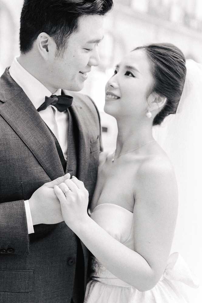 A black-and-white close-up portrait of a couple during what appears to be a wedding portrait session, photographed outdoors with a softly blurred architectural background. The bride wears a strapless gown and a veil, with her hair in a sleek updo, pearl drop earrings, and a delicate necklace; she looks up at the groom with a smile. The groom wears a fitted three-piece suit with a bow tie and holds the bride's hands, which show a ring on her finger. The image is shot with high contrast and bright highlights, giving it a clean, classic feel, with the couple positioned in a slightly angled portrait composition from roughly chest height upward.
