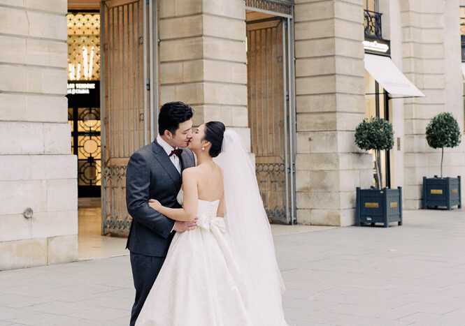 A couple portrait shot outdoors in front of a classical Haussmann-style stone building facade, likely Place Vendôme in Paris. The bride and groom are sharing a kiss, with the groom's hands around the bride's waist; he is wearing a charcoal grey suit with a burgundy bow tie, while the bride wears a strapless ivory ball gown with a large satin bow at the waist and a long cathedral-length veil. The image is a medium portrait composition with soft, even daylight, and topiary trees in dark planters are visible in the background alongside ornate doorways and a glimpse of an illuminated interior.