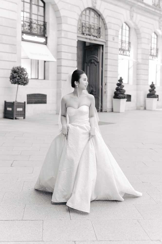 This black-and-white bridal portrait shows a bride standing alone on a stone-paved plaza in front of a grand Haussmann-style building with arched windows, ornate ironwork balconies, and a large wooden entrance door. The bride wears a strapless sweetheart-neckline ball gown with a full skirt, which she holds out slightly with both hands as she gazes to one side. Her hair is pulled back sleekly and she wears drop earrings. The image has soft, bright tones with gentle contrast, emphasizing the volume of the gown against the pale stone facade. The composition is a full-length portrait shot with the architectural backdrop slightly out of focus.
