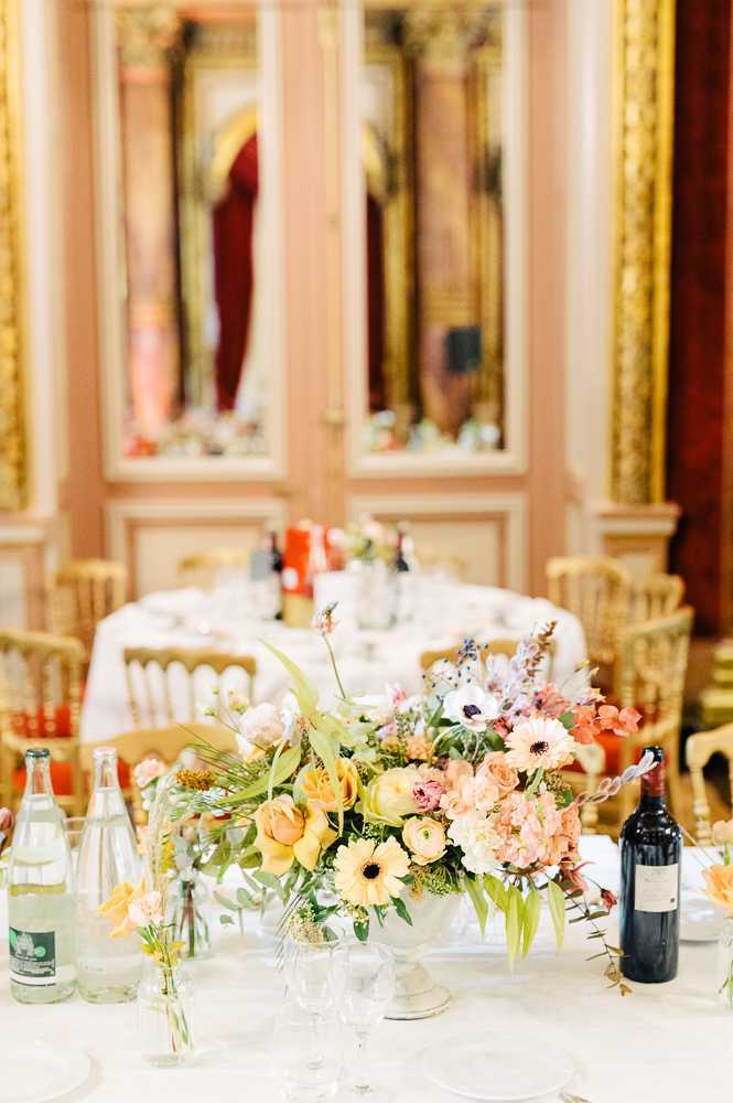 A reception table detail shot taken inside an ornate ballroom with blush pink walls, gold gilded moldings, and large gold-framed mirrors in the background. The foreground centerpiece is a lush, garden-style arrangement in a white footed compote vase, featuring yellow ranunculus, peach roses, white anemones with dark centers, blush carnations, soft pink blooms, lavender sprigs, and trailing greenery. The table is set with white linens, crystal glassware, water bottles, a red wine bottle, and small bud vases with single stems. In the background, additional round tables with white linens and gold Chiavari chairs are visible, with orange candles as accents, suggesting a classic French Parisian interior reception setting. The overall decor palette is warm peach, yellow, and blush with gold accents throughout the room.