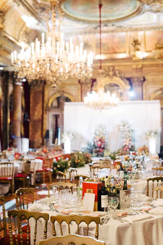 Wedding reception setup shot inside a grand Haussmann-era Parisian ballroom featuring ornate gilded architecture, painted ceiling murals, and large crystal chandeliers with candle-style lighting. The foreground shows a round reception table dressed in a white linen cloth, set with gold Chiavari chairs, crystal glassware, silverware, wine bottles, water carafes, and a red box centerpiece with mixed floral accents. In the background, multiple similarly dressed tables are visible alongside a white draped backdrop adorned with an abundant floral installation in blush, coral, and ivory tones. The overall decor palette combines white linens, gold chairs, and warm floral tones against the venue's ornate architectural details. Wide interior shot with shallow depth of field emphasizing the foreground table. Potential venue feature image.