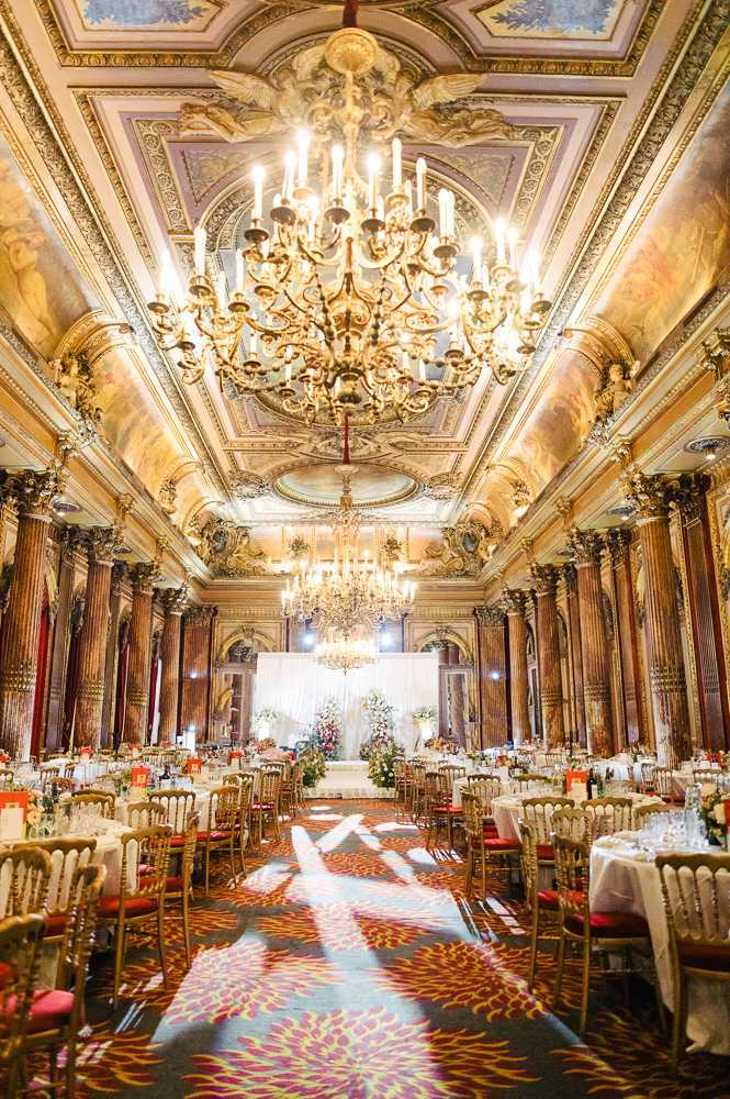 Wide interior shot of a grand ballroom set up for a wedding reception, with no guests present. The room features ornate gilded ceiling moldings with painted fresco panels, tall marble columns lining both sides, and two large gold candelabra-style chandeliers illuminating the space. Round reception tables dressed in white linen are arranged on either side of a central aisle, with gold Chiavari chairs featuring red cushions. The boldly patterned carpet runs in deep reds, oranges, and yellows with a floral motif. At the far end, a white draped backdrop with large floral arrangements in whites and pinks serves as a focal altar or head table display. The decor palette combines gold, white, and red accents. Potential venue feature image.