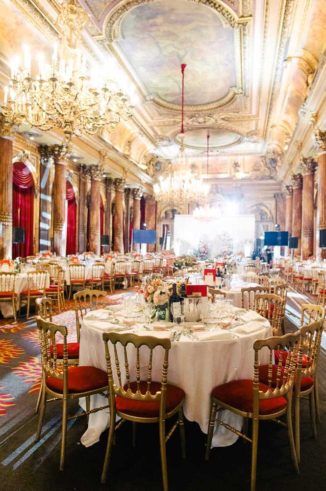 Wide interior shot of a grand ballroom reception setup with no guests present, showing the room dressed and ready for a wedding dinner. The space features ornate gilded architecture with tall marble columns, deep red draped curtains, and a painted ceiling fresco surrounded by gold molding, all lit by multiple large gold crystal chandeliers. Round tables are dressed in white linen and set with glassware, silverware, and small centerpieces of blush and peach flowers, with gold Chiavari-style chairs featuring red cushioned seats arranged around each table. A floral installation or backdrop is visible at the far end of the room near a bright light source, and a patterned carpet in red, gold, and blue runs throughout the floor. Potential venue feature image.