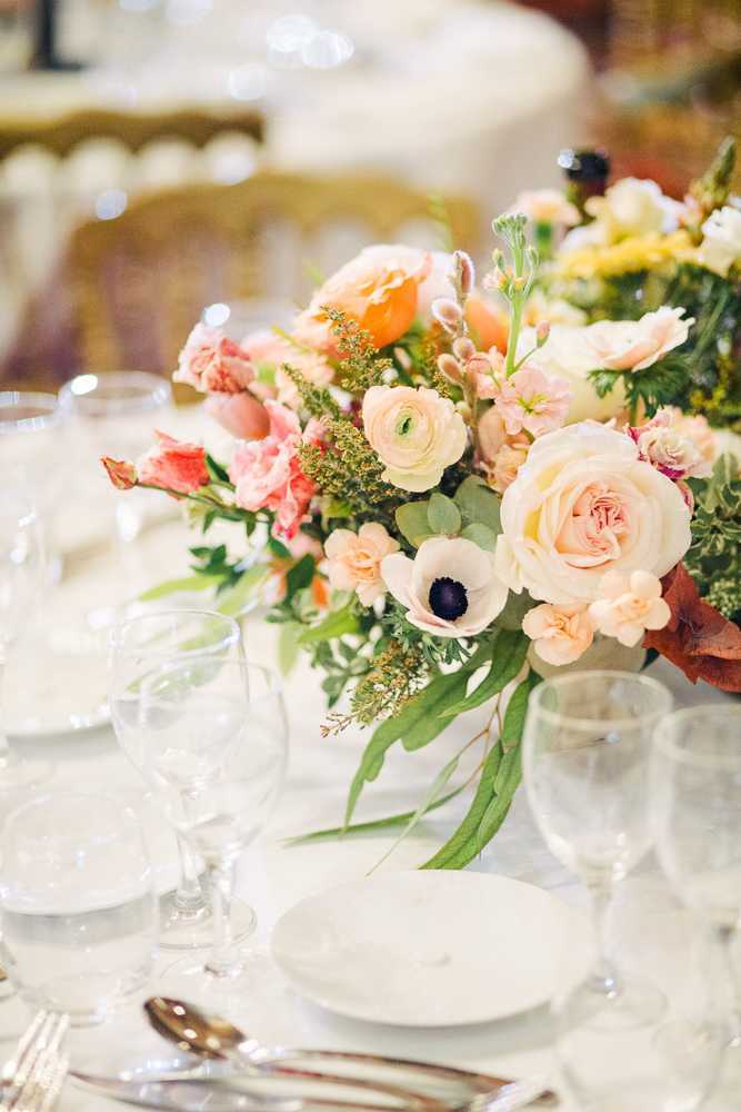 Close-up detail shot of a reception table centerpiece arrangement in an indoor ballroom setting, with gold chiavari chairs visible in the soft-focus background. The low centerpiece features a lush mix of cream garden roses, white anemones with dark centers, peach ranunculus, coral lisianthus, orange blooms, yellow filler flowers, and mixed greenery including eucalyptus, arranged in a white vessel. The table is set with white linens, white porcelain plates, clear crystal stemware, and silver cutlery, suggesting a classic, garden-inspired reception aesthetic with a warm peach, coral, and cream color palette.